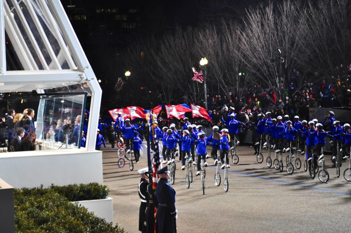 Gym Dandies riding into a bright spotlight at night. President Obama looks on from behind a protective glass structure